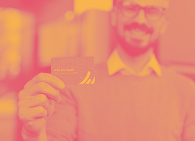 Foto: Ein lächelnder Mann mit Bart und Brille hält seine AVS Abo-Card in die Kamera. Darüber liegt ein Farbfilter in Gelb und Pink.