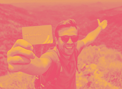 Foto: Ein junger Mann mit Sonnenbrille und Rucksack steht lachend auf einem Berg und hält die AVS Tourist-Card in die Kamera. Darüber liegt ein Farbfilter in Pink und Gelb.