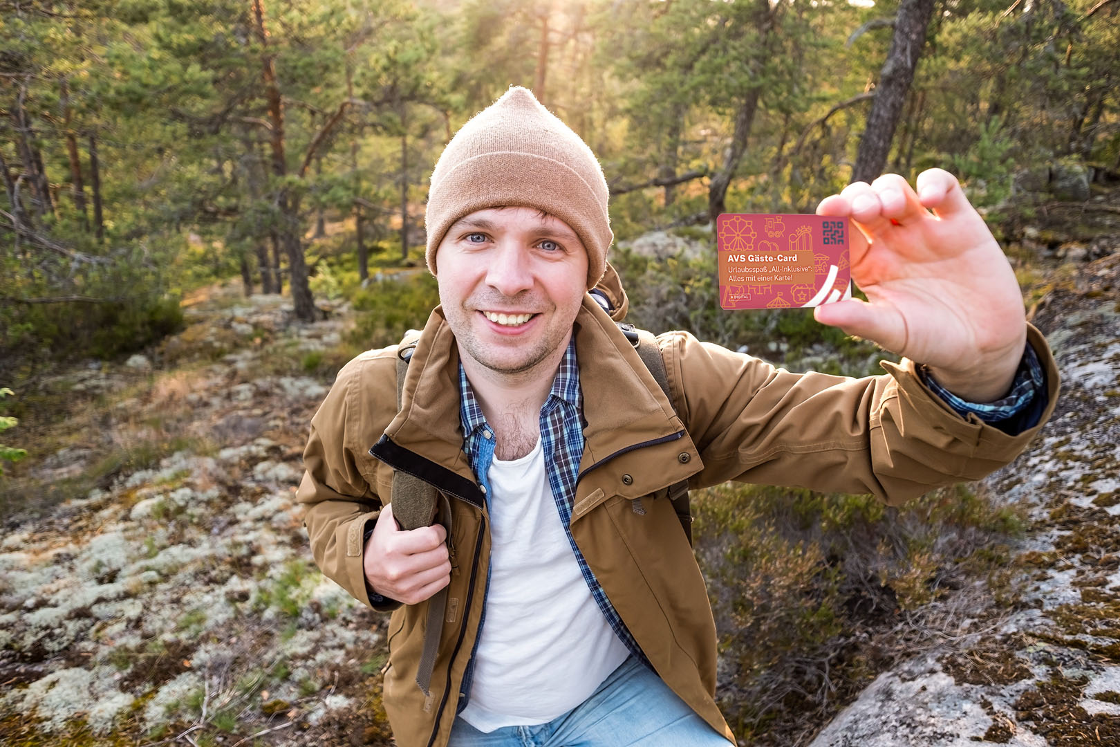 Foto: Ein junger Wanderer steht im Wald und hält lächelnd die AVS Gäste-Card in die Kamera.